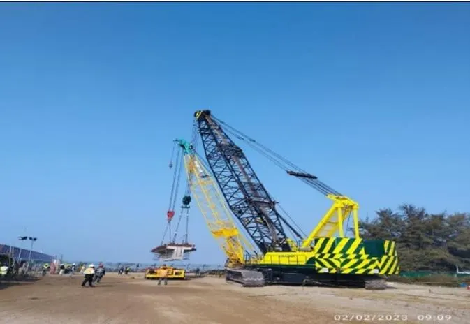 A yellow crawler crane with its lattice boom raised on a coastal project site.